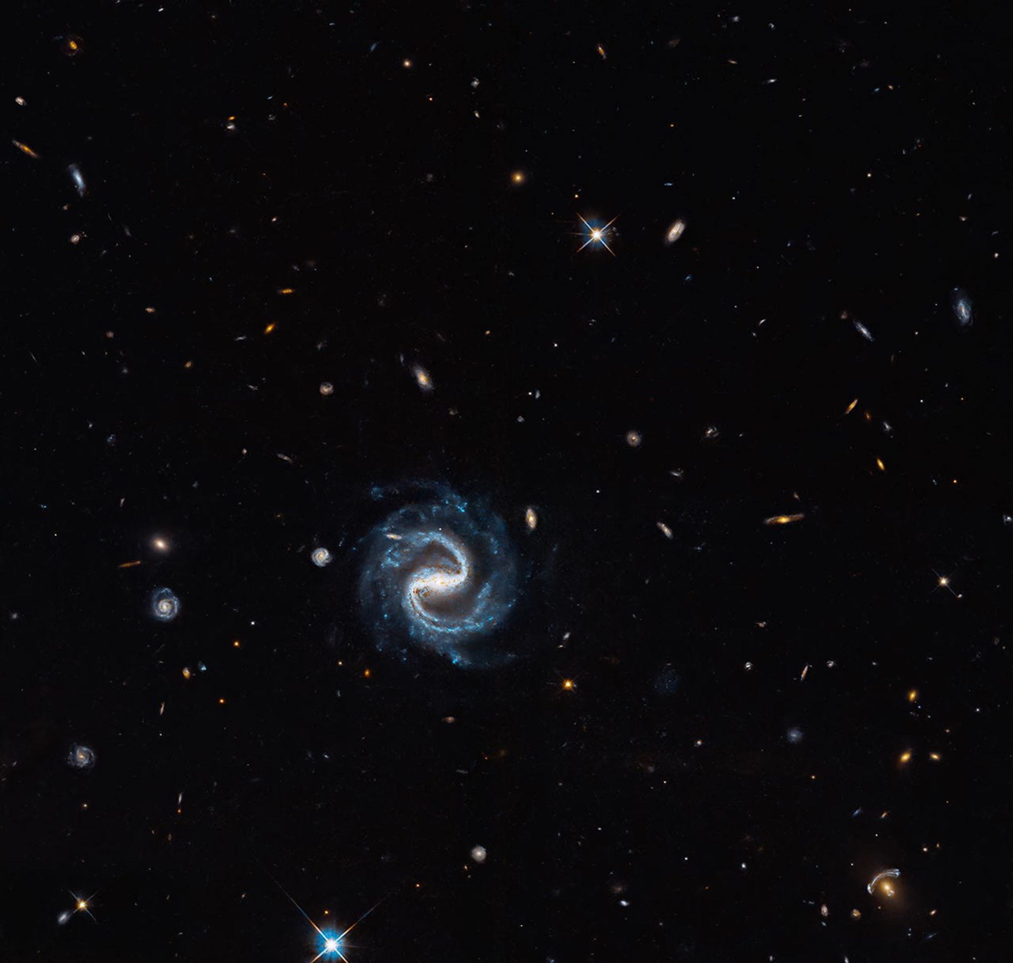 A barred spiral galaxy and a blue gravitationally lensed arc appear among distant galaxies.